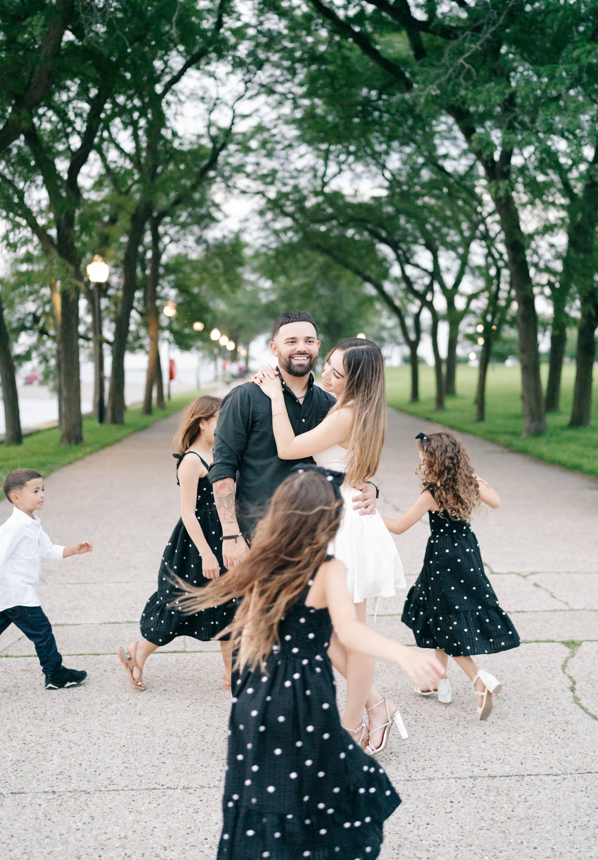 Parents embracing on a tree-lined lakefront promenade while their four children run and dance around them